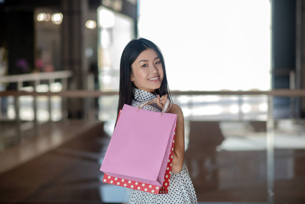 Happy brunetka kobieta niesie torby na zakupy na jednym ramieniu, uśmiechnięty - Zdjęcie, obraz