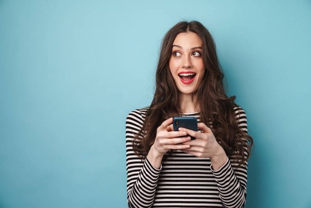 Foto van opgewonden mooie vrouw in gestreepte trui glimlachen en met behulp van mobiele telefoon geïsoleerd over blauwe muur - Foto, afbeelding