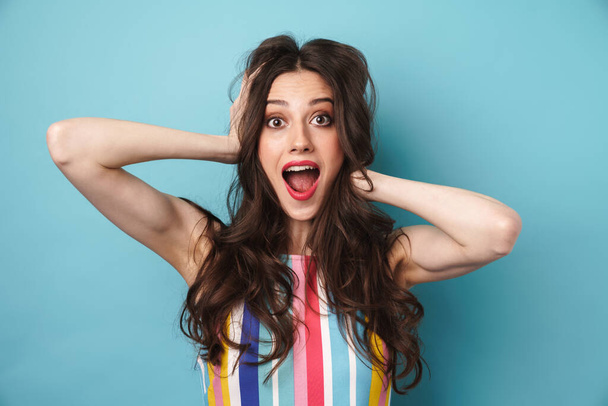 Photo of excited nice woman in striped dress grabbing her head and expressing surprise isolated over blue wall - Zdjęcie, obraz
