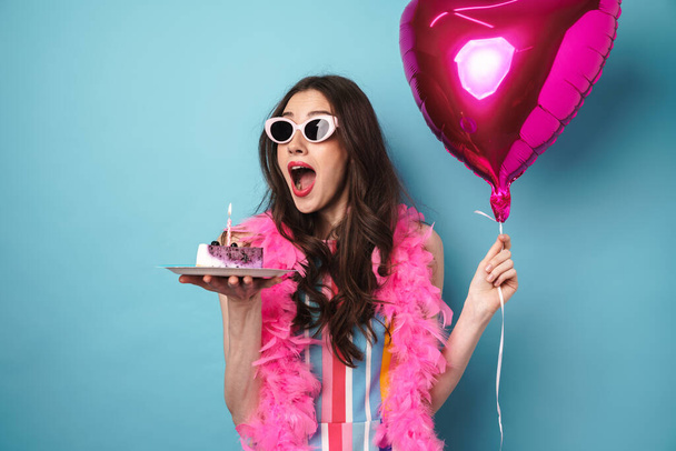 Photo of delighted young woman in sunglasses posing with balloon and cake isolated over blue wall - Foto, Imagem