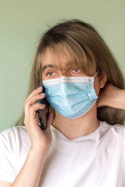 young woman with blond hair wearing a blue face mask and a white t-shirt calling through her gray mobile phone looking outside worried in a room - Photo, Image