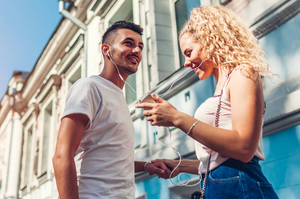 Szczęśliwa mieszana para wyścigowa zakochana w miłości słuchając muzyki przez telefon i tańcząc na ulicy miasta za pomocą słuchawek. Młodzi ludzie dobrze się bawią - Zdjęcie, obraz