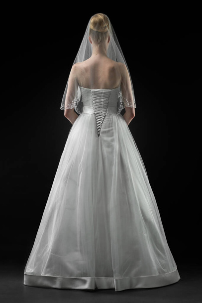 Bride in a beaituful long white wedding dress and lace bridal veil, back view. Black background - Foto, Imagen