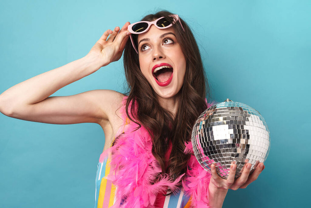 Photo of joyful young woman in sunglasses smiling while posing with disco ball isolated over blue wall - Valokuva, kuva