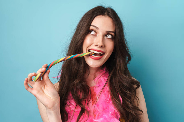Fotografie radostné krásné ženy v peří boa usmívat a jíst sladkosti izolované přes modrou zeď - Fotografie, Obrázek
