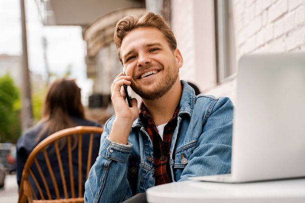 Красивый улыбающийся счастливый молодой хипстер в уличном кафе, разговаривающий по мобильному телефону во время работы над ноутбуком. Современные технологии, онлайн связь и интернет. Люди образ жизни
 - Фото, изображение