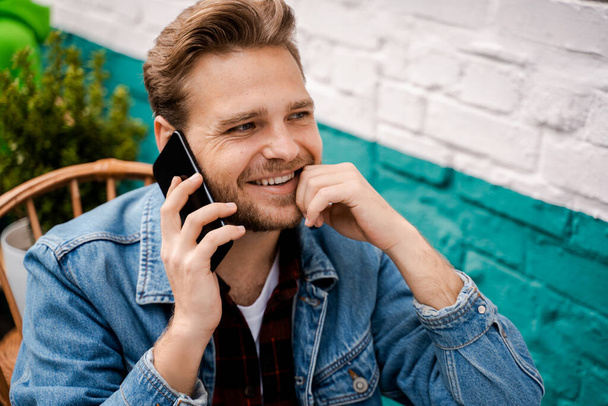Sorrindo bonito barbudo jovem com um corte de cabelo elegante fala ao telefone enquanto sentado fora na rua em um café, pensa e sonha, agradável conversa relaxada. Estilo de vida das pessoas
 - Foto, Imagem