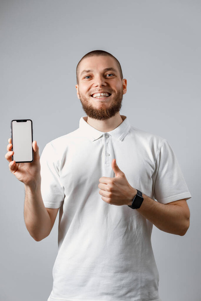 灰色の背景に電話画面を示す白いTシャツの幸せな若い男の肖像画。隔離された。コピースペース - 写真・画像