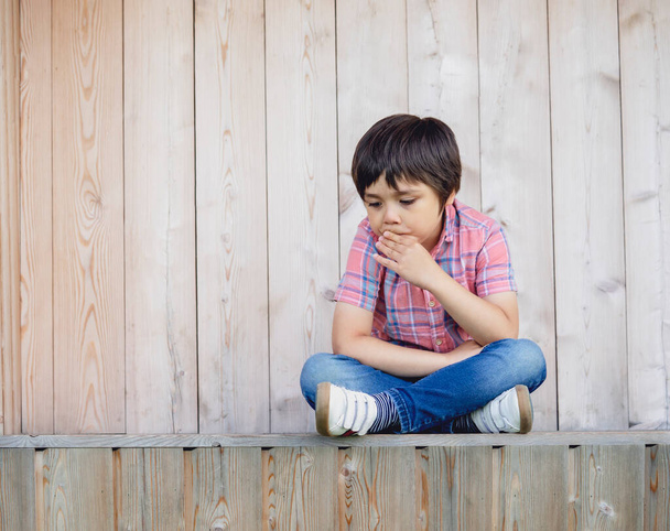 Emotional portrait of Unhappy boy sitting next to wooden wall with sad face, Upset kid sitting alone and looking down with thinking face, Lonely child with bored face,Mental health child concept - Photo, Image