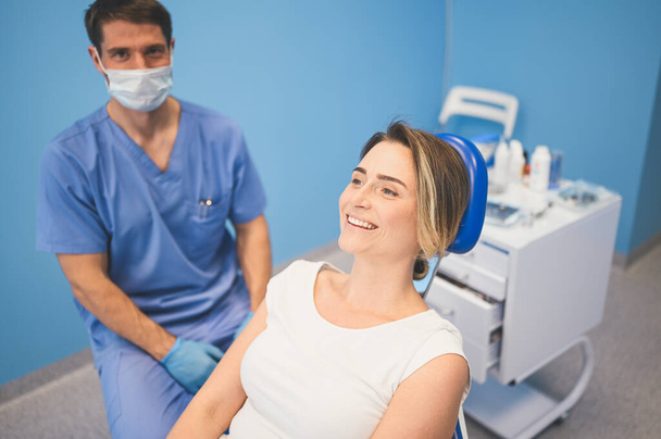Dentist examining a patient's teeth using dental equipment in dentistry office. Stomatology and health care concept. Young handsome male doctor in disposable medical facial mask, smiling happy woman. - Photo, Image