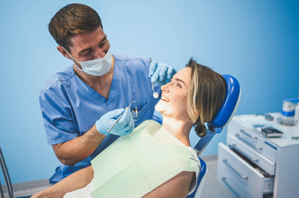 Fogorvos, aki fogászati felszereléssel vizsgálja a páciens fogait a fogorvosi rendelőben. Fogászati és egészségügyi koncepció. Fiatal jóképű férfi orvos eldobható orvosi arc maszk, mosolygós boldog nő. - Fotó, kép