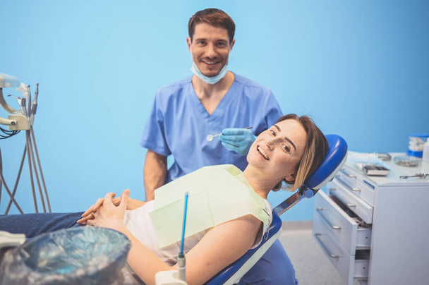 Diş hekimi muayenehanesinde bir hastanın dişlerini diş teçhizatı kullanarak muayene ediyor. Stomatoloji ve sağlık hizmetleri kavramı. Tek kullanımlık tıbbi maskeli yakışıklı erkek doktor, gülümseyen mutlu kadın.. - Fotoğraf, Görsel