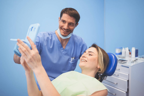 Zubař ukazuje pacientovi výsledky léčby zrcadlem, při prohlídce zubů zubním nářadím v zubní ordinaci. Mladý pohledný mužský lékař v lékařské masce obličeje a usmívající se šťastná žena. - Fotografie, Obrázek