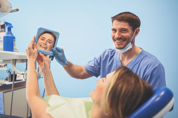 Le dentiste montre au patient les résultats du traitement avec un miroir, examinant les dents avec du matériel dentaire dans le cabinet dentaire. Jeune beau médecin masculin en masque facial médical et sourire femme heureuse
. - Photo, image