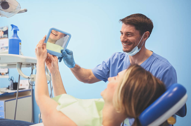 El dentista muestra al paciente los resultados del tratamiento con un espejo, examinando los dientes con equipo dental en el consultorio dental. Joven guapo médico masculino en máscara facial médica y sonriente mujer feliz
. - Foto, imagen