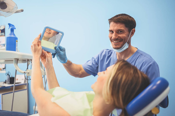 El dentista muestra al paciente los resultados del tratamiento con un espejo, examinando los dientes con equipo dental en el consultorio dental. Joven guapo médico masculino en máscara facial médica y sonriente mujer feliz
. - Foto, Imagen