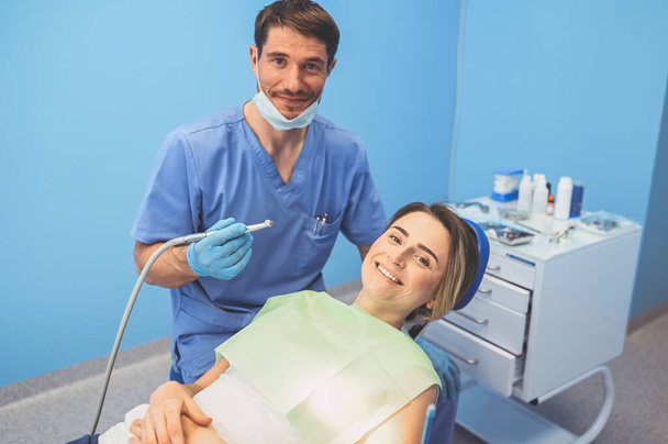 Dentista examinando los dientes de un paciente usando equipo dental en la oficina de odontología. Concepto estomatológico y asistencial. Joven guapo médico varón en mascarilla facial médica desechable, sonriendo mujer feliz
. - Foto, Imagen