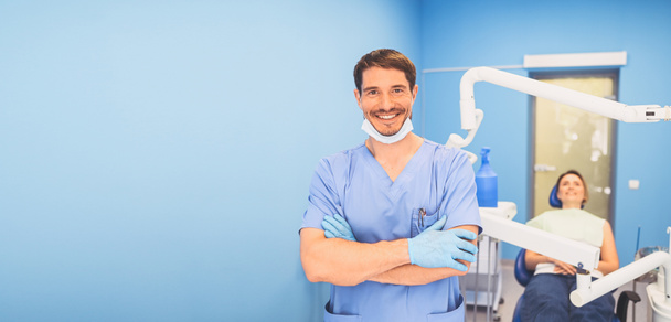 Jovem sorridente bonito médico masculino em azul uniforme médico, descartável máscara facial médica com equipamentos no consultório odontológico e paciente feliz na cadeira dentária. Conceito de estomatologia. Espaço de cópia
 - Foto, Imagem