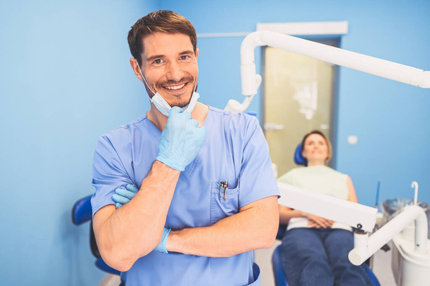 Jonge lachende knappe mannelijke arts in blauw medisch uniform, wegwerp medisch gezichtsmasker met apparatuur in de tandheelkunde kantoor en gelukkige patiënt in de tandartsstoel. Stomatologie concept. Kopieerruimte - Foto, afbeelding