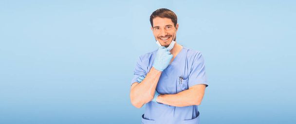 Jonge lachende knappe mannelijke tandarts in blauw medisch uniform, wegwerp medisch gezichtsmasker en beschermende handschoenen op de blauwe achtergrond geïsoleerd. Dental Stomatologie concept. Kopieerruimte - Foto, afbeelding