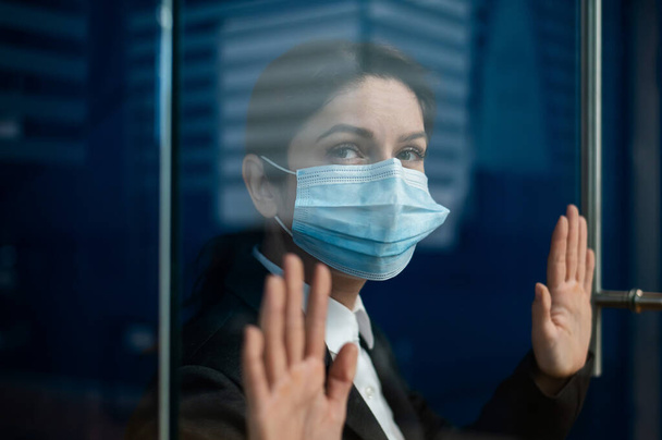 A sad quarantined woman in a business suit leaned against the window. Caucasian girl in a mask on isolation put her hands on a glass door. Maintaining social distance to prevent coronavirus. - Foto, Imagen