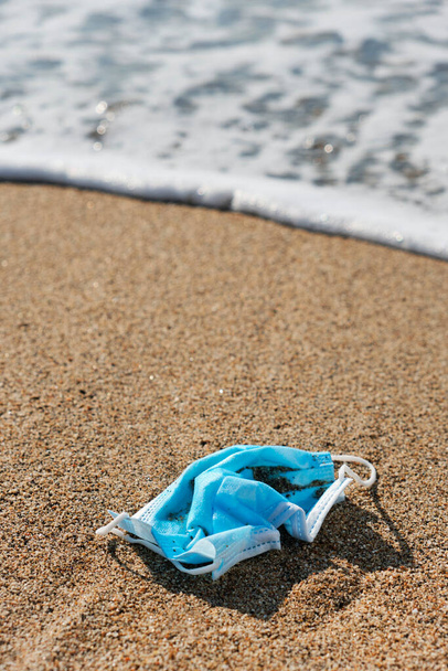 gros plan d'un masque chirurgical bleu jeté sur le sable mouillé du bord de mer d'une plage - Photo, image