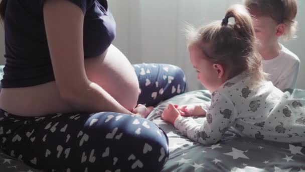 Pregnancy, motherhood, family, childhood, upbringing, training, self-isolation concept - young pregnant girl with small children on bed. Girl and boy caress and kiss whole belly of mom against window. - Video