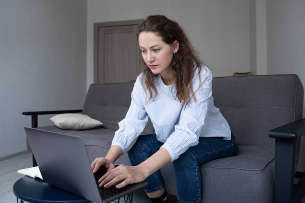 A girl in a blue shirt and jeans is working on a laptop sitting on the sofa. Working environment at home - Foto, Imagen