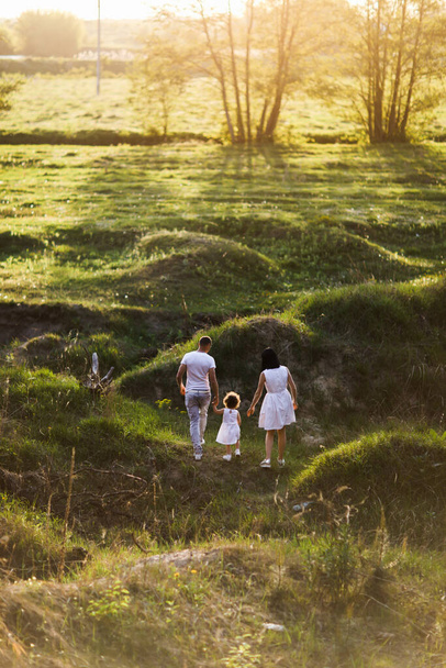Çimlerle kaplı yamaçlarda çocuklu bir çift yürüyor. Gün batımı. Dikey fotoğraf. - Fotoğraf, Görsel