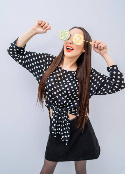 Beautiful brunette girl holds two lollipops near eyes. Cheerful lady in dotted shirt over white background. - Valokuva, kuva