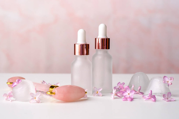 Cosmetic bottles of essential oil, face massage roller and lilac flowers on pink background. Aromatherapy, facial skin care concept. - Foto, immagini