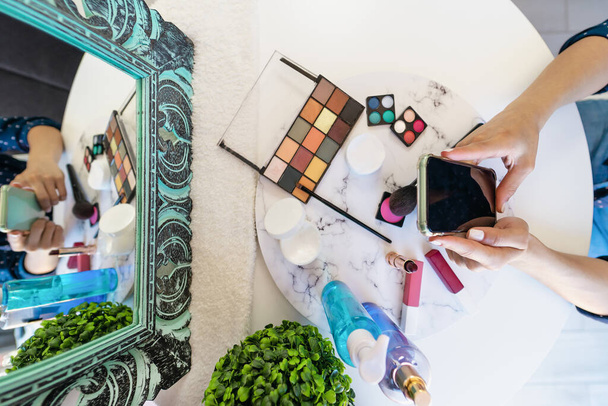Top view νεαρή γυναίκα χρησιμοποιώντας κινητό smartphone, ενώ κάνει μακιγιάζ στο σπίτι - Αισθητική Ομορφιά βιομηχανία και την τεχνολογία εξαρτημένοι άνθρωποι έννοια - Φωτογραφία, εικόνα