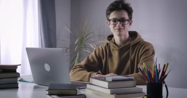 Portrait of nerd Caucasian college student sitting at the table with laptop and books, and looking at camera. Smiling happy brunette boy in eyeglasses posing indoors. Cinema 4k ProRes HQ. - Felvétel, videó