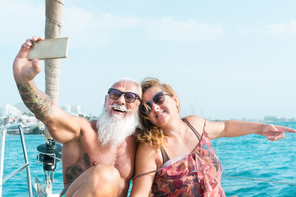 Senior couple taking selfie with mobile smartphone on sailboat vacation - Happy mature people having fun celebrating wedding anniversary on boat trip - Love relationship and travel lifestyle concept - Photo, Image