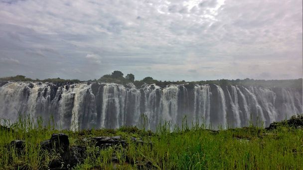 Egyedülálló Victoria Falls. A Zambezi folyó számos hatalmas vízfolyást hoz le a szurdok szélén keresztül. Az előtérben van egy másik széle, amelyet élénk zöld fű borít. Az ég felhős.. - Fotó, kép