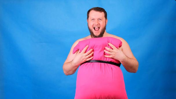 枕で作られたピンクのドレスを着た面白い髭の男が青い背景に胸に触れる。狂気の隔離だ。面白い家の掃除。ファッション2020 。枕の上に置け。課題2020年の住宅分離による - 写真・画像