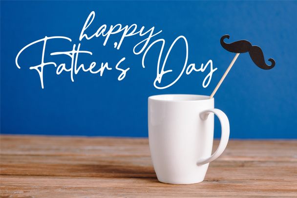 white cup and black decorative paper fake mustache on wooden surface isolated on blue, happy fathers day illustration - Photo, Image