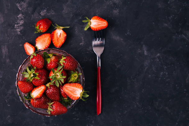 One strawberry on a red fork and in the crystal bowl of the black rustic background. Top view. Vertical. Diet minimal concept. - Φωτογραφία, εικόνα