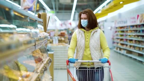 Young woman in medical mask and gloves by shelves with fresh pastry at store - Felvétel, videó