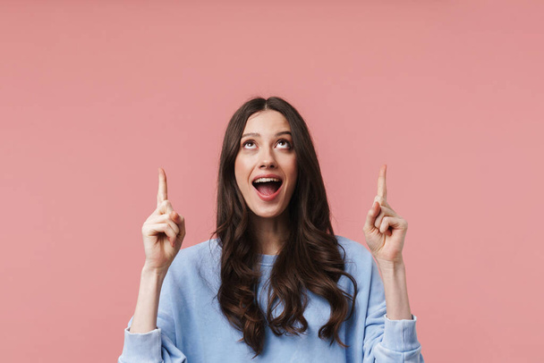 Image of attractive young woman with long brown hair smiling and pointing fingers at empty space isolated over pink background - Foto, afbeelding