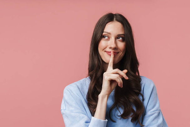 Image of attractive young woman with long brown hair showing hush gesture while keeping finger at lips isolated over pink background - Fotografie, Obrázek