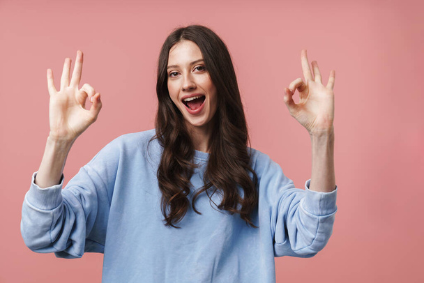 Image of attractive young woman with long brown hair smiling and gesturing okay sign isolated over pink background - Foto, afbeelding