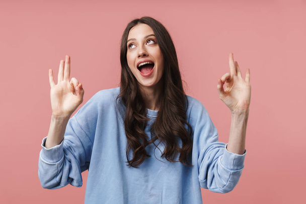 Image of attractive young woman with long brown hair smiling and gesturing okay sign isolated over pink background - Фото, изображение