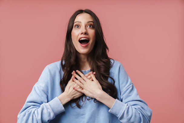 Image of attractive young woman with long brown hair screaming in delight and holding hands at her chest isolated over pink background - Foto, Bild