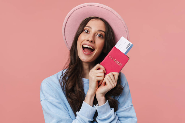 Portrait of a pretty cheerful girl with long curly brunette hair wearing sweatshirt standing isolated over pink background, showing passport with flight tickets - Photo, Image
