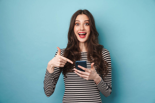 Фото взволнованной милой женщины в полосатом свитере, улыбающейся и указывающей пальцем на мобильный телефон, изолированный над синей стеной
 - Фото, изображение