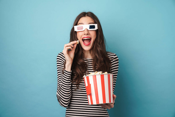 Photo of excited young woman in 3d glasses eating popcorn while watching movie isolated over blue wall - Fotografie, Obrázek