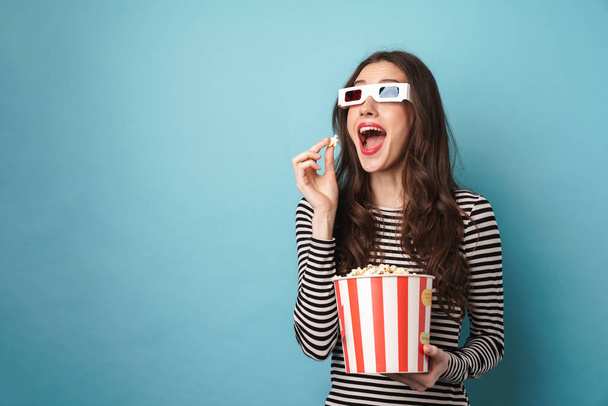 Photo of excited young woman in 3d glasses eating popcorn while watching movie isolated over blue wall - Fotó, kép