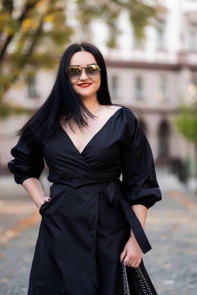 Autumn fashion outdoor. The girl with red lips in fashionable stylish black dress and sunglasses, autumnal lifestyle on the background of blurry yellow-green trees in the park. Vertical - Fotó, kép