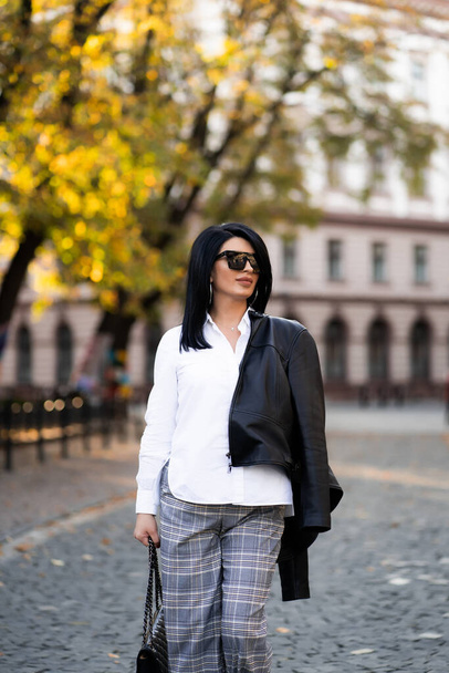 Autumn fashion outdoor. The girl in fashionable stylish white shirt , pants, jacket and sunglasses, autumnal lifestyle on the background of blurry yellow-green trees in the park. Vertical - Photo, Image